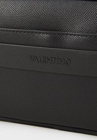 A bolsa de couro preto texturizado apresenta um logótipo em relevo proeminente e uma alça de accentuamento suave para um design estruturado.