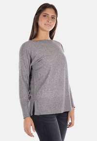 Maglione grigio lavorato a maglia con maniche lunghe, scollatura rotonda, fessure ai lati dell'orlo e una texture morbida. Indossato sopra jeans scuri.
