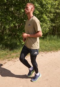 Mężczyzna biegający po ścieżce z ziemi obok zielonych drzew, ubrany w beżową koszulę Nike Trail, czarne spodenki na legginsy i szare oraz zielone buty do biegania.