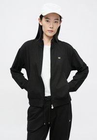 Melnā zipa jaka ar augstu apkakli, izgatavota no mīksta auduma, valkāta kopā ar baltu t-kreklu zemāk. Zelta akcenti uz metāla detaļām, minimālistisks dizains.