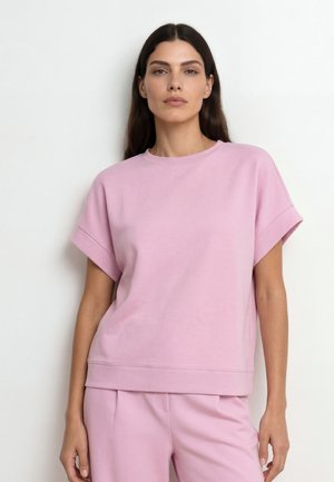 Nő laza szabású, rövid ujjú pasztell rózsaszín felsőben és hozzáillő nadrágban, egyszínű fehér háttér előtt áll.