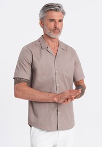 Beige Kurzarmshirt aus glattem Stoff, mit Reverskragen, vorderem Knopfverschluss und geradem Saum, kombiniert mit schlichten weißen Hosen.