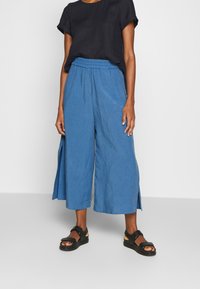 Blauwe, wijde culottebroek met zijsplitten, gemaakt van lichtgewicht stof. Draagt een zwart, kort mouwen top en zwarte sandalen.