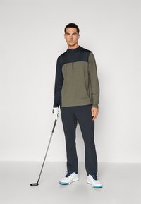 Herre golf antrekk med en mørkegrønn og marineblå quarter-zip genser, mørke bukser, hvite sko med blå detaljer, og en golfkølle.