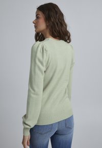 Lys grøn sweater med struktureret strik, puffede skuldre og ribbede kanter og kant, parret med blå denimjeans.