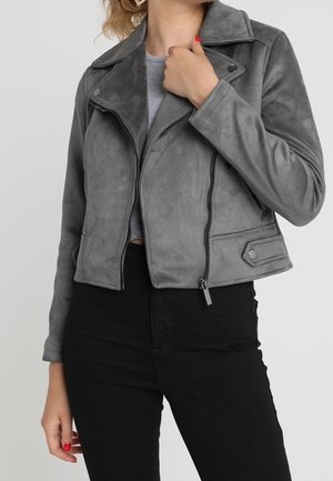 Femme portant une veste de motard en daim gris par-dessus un haut gris et un pantalon noir, tenant le col de la veste d'une main.