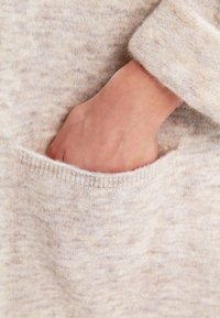 Helles beige Strickpullover mit einer Tasche, in der eine sichtbare Hand platziert ist. Die Textur erscheint weich und leicht fusselig.