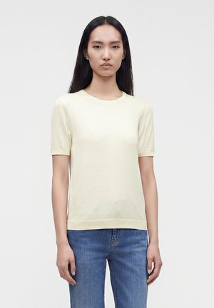 Kvinde med langt mørkt hår iført en lys cremefarvet kortærmet sweater og blå jeans, der står foran en ensfarvet grå baggrund.