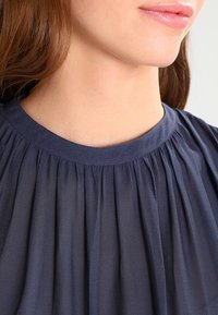 Kvinnas nedre ansikte och hals som bär en navyblå, veckad högkragsblus med lång brunt hår synligt.