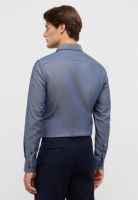 Herenoverhemd in een donkerblauwe, gestructureerde stof. Heeft lange mouwen, een nette kraag en knoopmanchetten. Gedragen met donkere broeken.