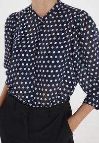 Blouse en bleu marine, transparente, à pois blancs, avec un col à boutons et des manches courtes bouffantes, portée avec un pantalon noir.