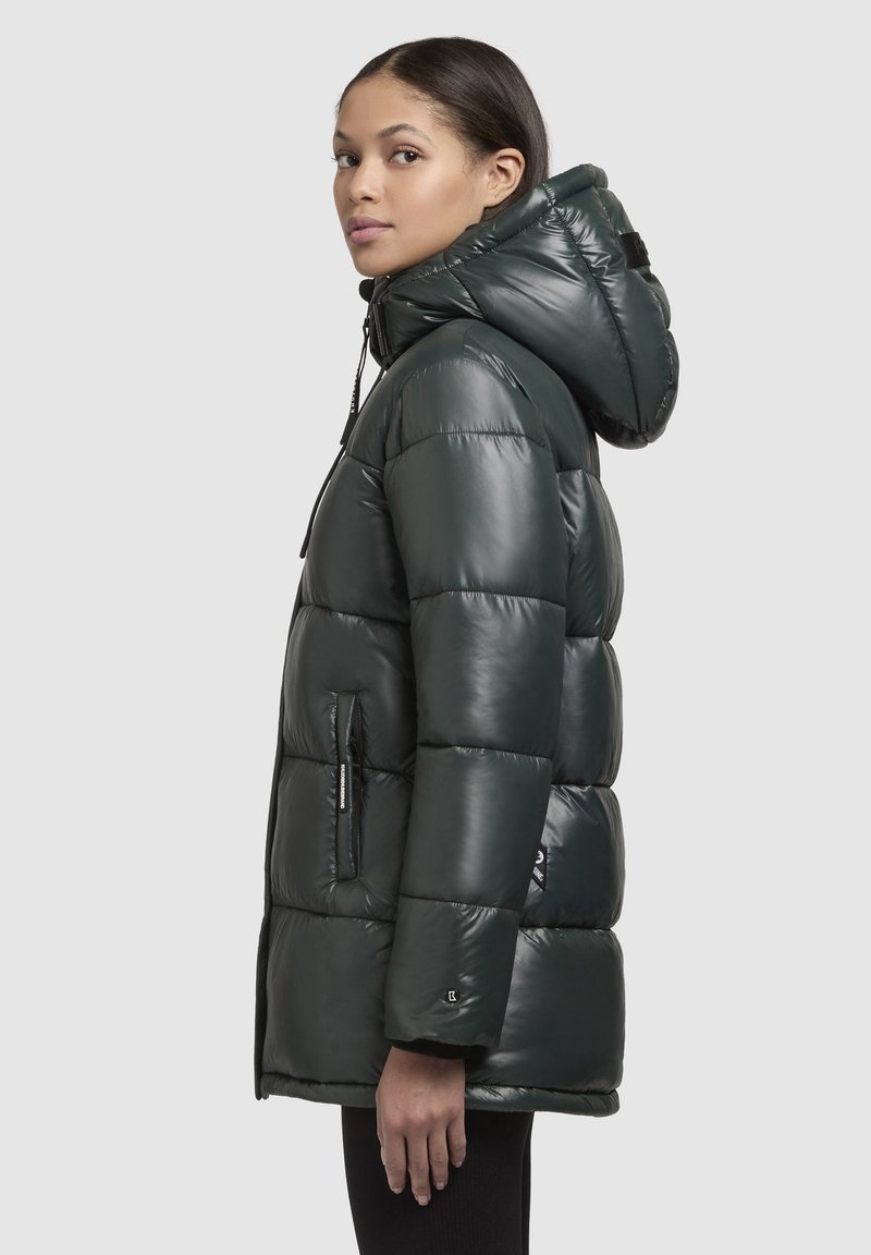 Giacca puffer verde scuro con cappuccio, caratterizzata da sezioni trapuntate e tasche con zip. Realizzata in tessuto lucido e liscio con dettagli a cordoncino.