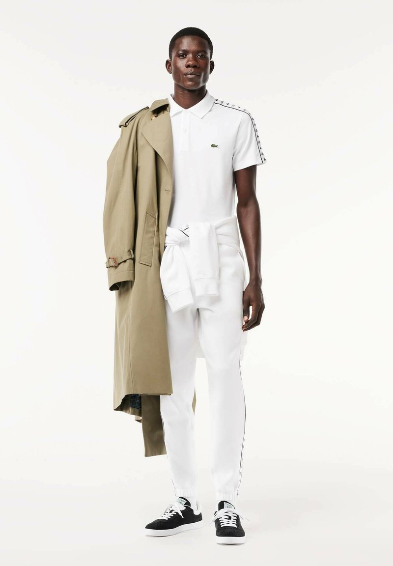 Polo blanc avec logo Lacoste, trench beige drapé sur l'épaule, joggers blancs, baskets noires avec semelles blanches, design épuré.
