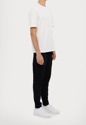 Mężczyzna ubrany w prostą białą koszulkę z krótkim rękawem, czarne spodnie i białe trampki, stojący w neutralnej pozie na jasnym tle.