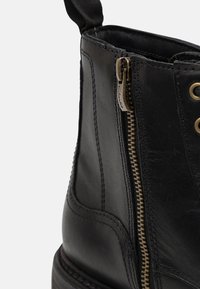 Botines de piel negra con cremallera lateral, detalles metálicos y costuras. El diseño incluye una lengüeta para facilitar su puesta.