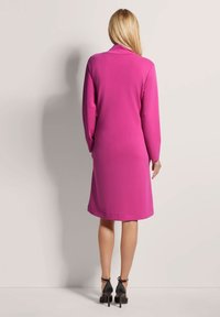 Langärmliges, knielanges Kleid aus leuchtend pinkem Stoff, glatte Textur, mit klassischem Kragen und geradem Schnitt. Kombiniert mit schwarzen High Heels.
