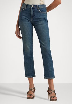 Kvinde iført korte mørkeblå jeans og brune højhælede sandaler, der står mod en ensfarvet baggrund.