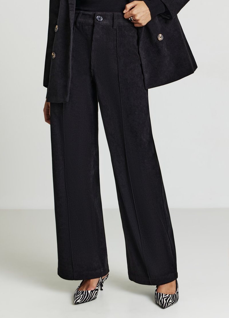 Schwarze Cordhose mit weitem Bein und strukturierter Oberfläche, mit hoher Taille und zwei vorderen Falten, kombiniert mit gemusterten Absatzschuhen.