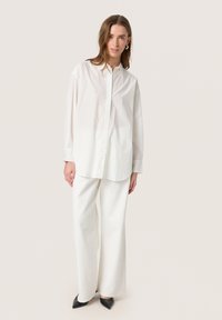 Cămașă albă cu guler, cu mâneci lungi, combinată cu pantaloni albi, cu textură, lejeri și pantofi negri cu vârful ascuțit. Linii simple și curate.