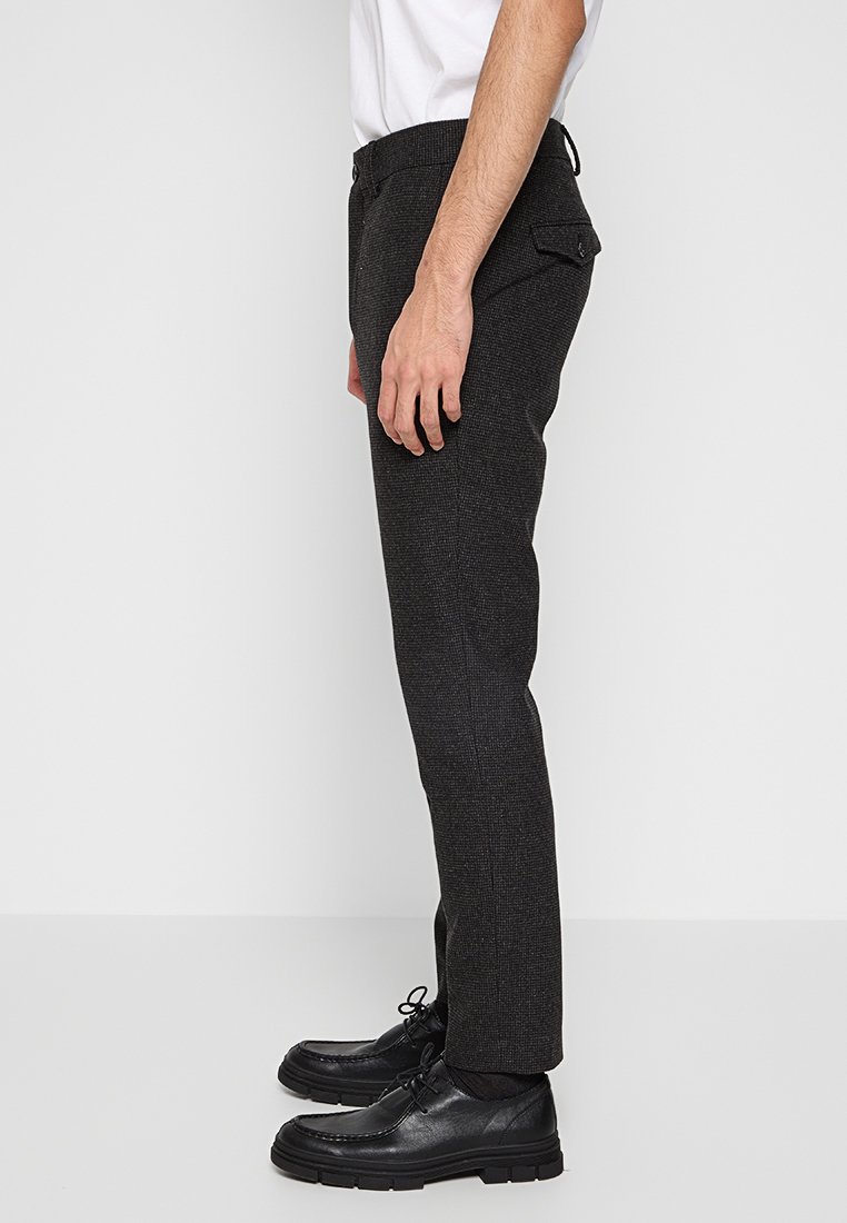 Maßgeschneiderte, dunkelgraue Hose aus strukturiertem Stoff, mit Seitentaschen und geradem Schnitt. Komplementiert durch schwarze Schnürschuhe.