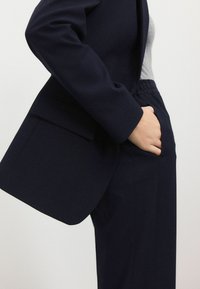 Námořnický modrý oblek se skládá z blejzru s elegantním límcem a šikmou kapsou, doplněný o kalhoty na míru. Hladká textura, moderní design.