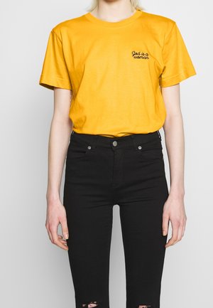 Geel katoenen T-shirt met korte mouwen, voorzien van zwart handgeschreven tekst "God is a woman" op de linkerborst, gecombineerd met zwarte gescheurde jeans.