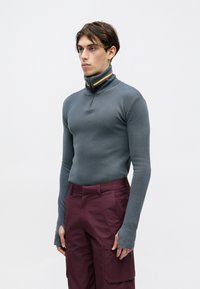 Mlad moški nosi prilegajočo se sivo rolko z zadrgo z mavričnimi črtami in bordo cargo hlačami, stoji pred enostavno belo ozadjem.