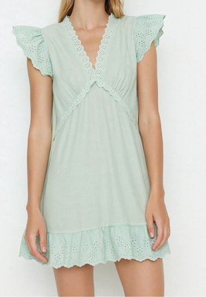 Mujer que lleva un vestido corto verde claro con ribete de encaje con bordes en forma de concha y mangas y bajo con volantes con ojales.