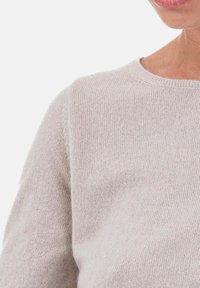 Gros plan d'une personne portant un pull en tricot beige clair, montrant l'épaule, le cou et une partie du visage sur un fond blanc.