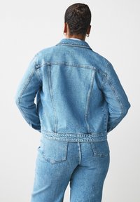 Lichtblauwe denimjack met een iets getailleerd ontwerp, zichtbare stiksels en een klassieke kraag, gecombineerd met bijpassende denimbroek.