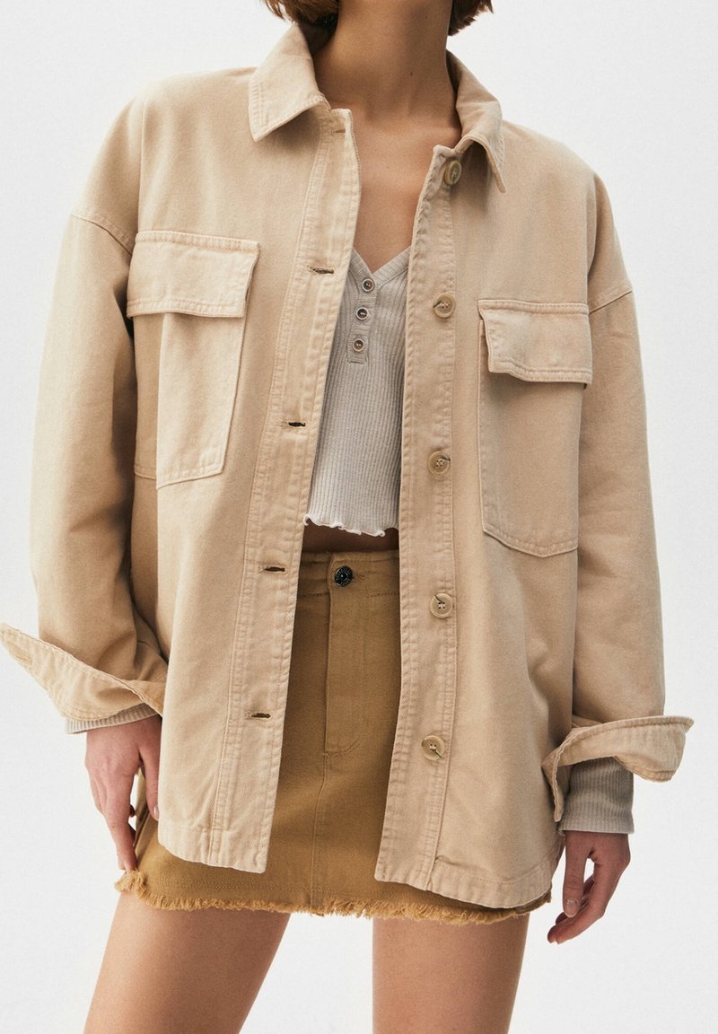 Veste en denim beige avec fermeture à boutons et deux poches avant, superposée sur un haut gris en rib à côtes, accompagnée d'une mini jupe beige à l'ourlet effiloché.
