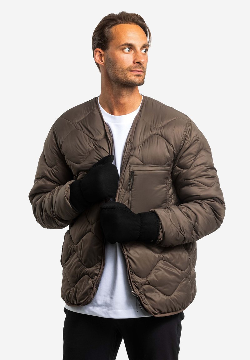 Gepolsterte braune Jacke mit Reißverschluss vorne, diamantgequilteter Textur und einer Brusttasche. Das Modell trägt schwarze Handschuhe und ein weißes T-Shirt.