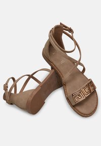 Hnědé semišové sandály s křížovými kotníkovými pásky, dekorativním zlatým geometrickým akcentem, plochou podrážkou a texturovanou gumovou podešví.