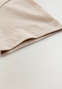 Tissu en coton beige clair plié, présentant une texture lisse et un ourlet cousu le long du bord. Une subtile élasticité est visible dans le matériau.