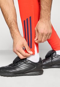 Pantalones deportivos rojos con detalles de tres rayas en azul marino en el lateral, combinados con zapatos deportivos negros y calcetines blancos. Cremallera visible en el dobladillo.