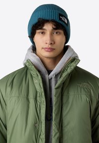 Grøn pufferjakke med sølvknapper, båret over en grå hættetrøje. Teal ribbet hue med et sort logo-patch foran. Neutral baggrund.
