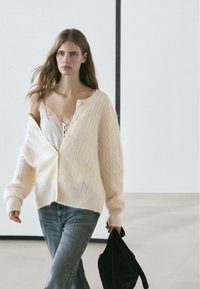 Cardigan a coste color crema con vestibilità comoda, indossato aperto sopra una canottiera bianca di pizzo, abbinato a jeans blu e una borsa nera.