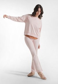 Sudadera rosa claro y pantalones a juego hechos de tela suave. El diseño incluye un cuello en v y un ligero acampanado en el dobladillo de los pantalones. Se observan detalles en marrón.