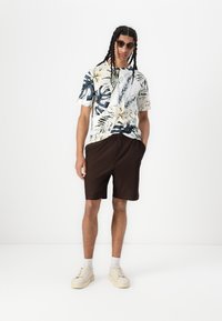 Camisa de manga corta con estampado floral en tonos verde oscuro y amarillo; pantalones cortos marrón a rayas; zapatillas blancas; gafas de sol oscuras.