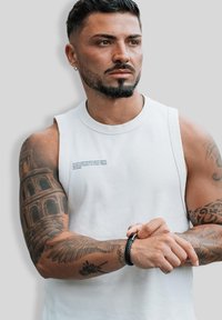 Man with short dark hair and beard wearing a sleeveless white shirt, showing detailed tattoos on both arms, adjusting a black wristband.
