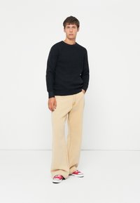 Giovane uomo in piedi con pantaloni beige a gamba larga, maglione nero testurizzato e sneakers rosse su uno sfondo bianco semplice.