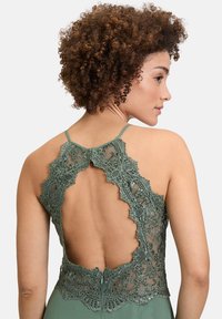 Zielona sukienka z koronkowym tyłem, ozdobiona skomplikowanymi wzorami kwiatowymi, z regulowanymi ramiączkami i wycięciem w kształcie dziurki, wykonana z gładkiego, swobodnie opadającego materiału.