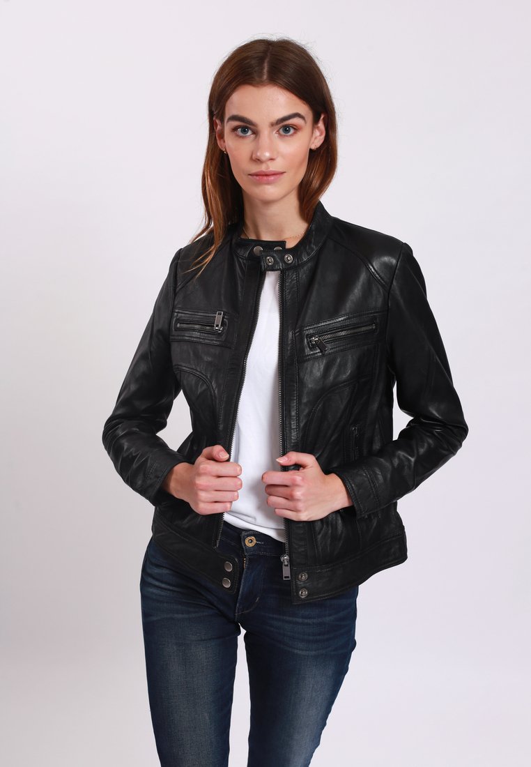 Giacca di pelle nera con colletto alto, tre tasche frontali con zip e design aderente. Indossata sopra una camicia bianca e abbinata a pantaloni jeans blu.