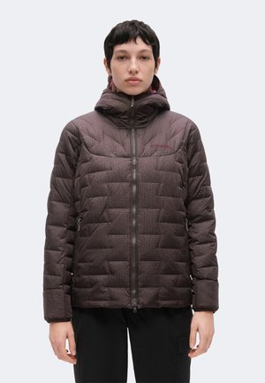 Persona con cabello corto y oscuro, que lleva una chaqueta acolchada con capucha de color marrón oscuro con cremallera frontal y pantalones negros sobre un fondo liso.