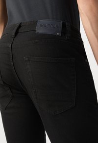 Jeans de mezclilla negra con una etiqueta de cuero que dice "INDICODE EST. 1991", con dos bolsillos traseros y detalles de costura sutiles.