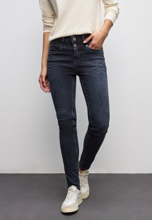 Dunkelblaue Skinny-Jeans mit hoher Taille, zwei-Knopf-Verschluss und Fronttaschen, kombiniert mit hellen High-Top-Sneakern.
