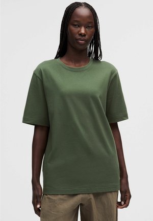 Vrouw met lange vlechten die een oversized olijfgroen T-shirt met korte mouwen en lichtbruine broek draagt, staand tegen een effen achtergrond.