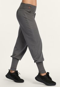 Eine Person trägt graue Jogginghosen und schwarze Sportschuhe, während sie in der Mitte des Schrittes vor einem schlichten hellen Hintergrund geht.