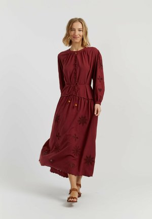 Donna che cammina con un vestito color bordeaux a maniche lunghe con bottoni frontali, ritagli floreali, vita con coulisse e sandali marroni su sfondo neutro.