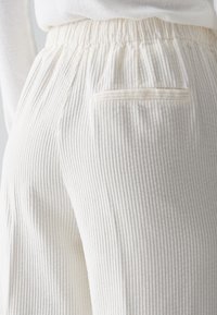 Pantalons en velours blanc avec une texture à côtes verticales, taille élastique et un détail de poche arrière, mettant en avant un tissu doux et confortable.